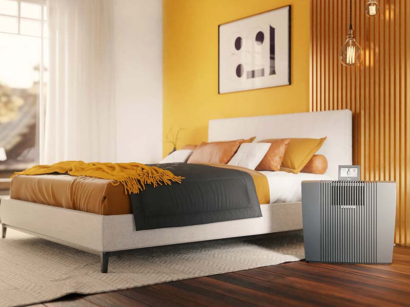 Venta Air Purifier Professional contra las alergias se alza en un dormitorio en tonos amarillos y naranjas.