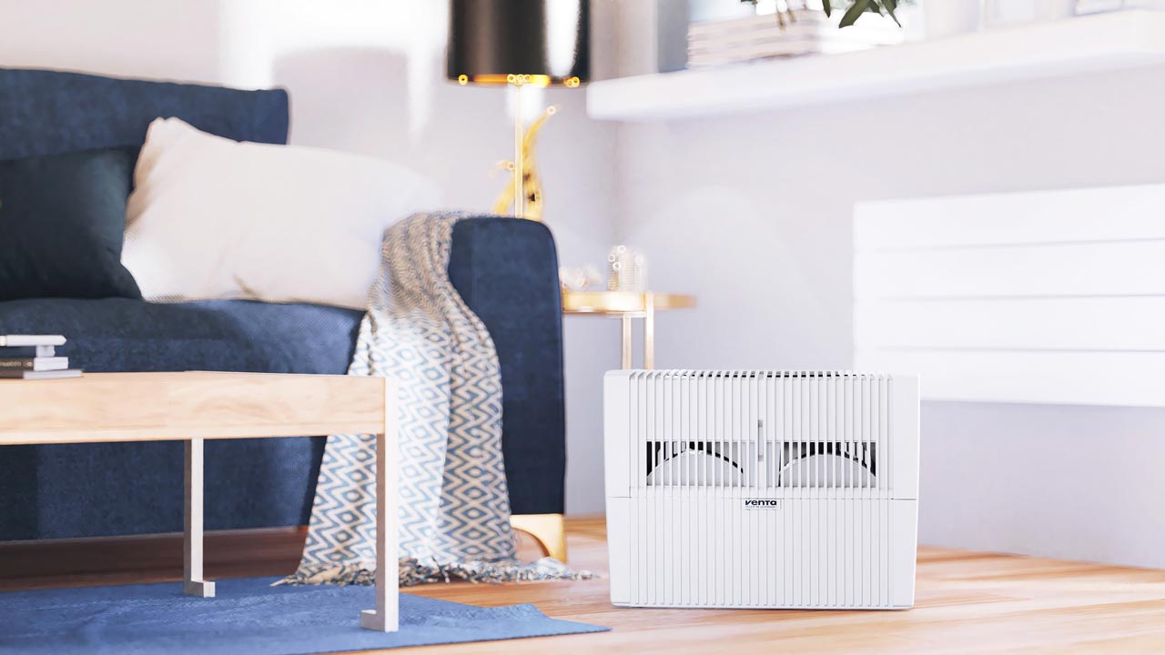Venta Luftbefeuchter Comfort Plus steht in einem Wohnzimmer mit blauer Couch, um dort für eine optimale Luftfeuchtigkeit von 40-60 Prozent zu sorgen.